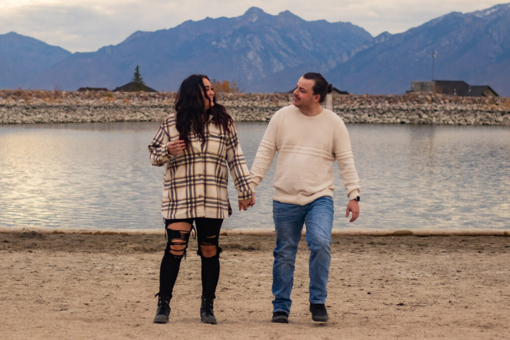 Couple walking towards the camera holding hands with the mountains behind them at Blackridge Reservoir in Herriman, UT