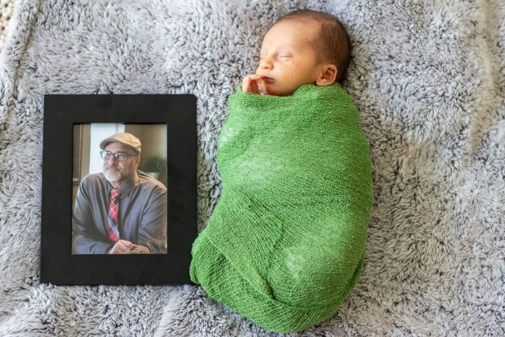 Newborn baby wrapped in a green swaddle laying on a blanket next to a framed photo of his late grandpa