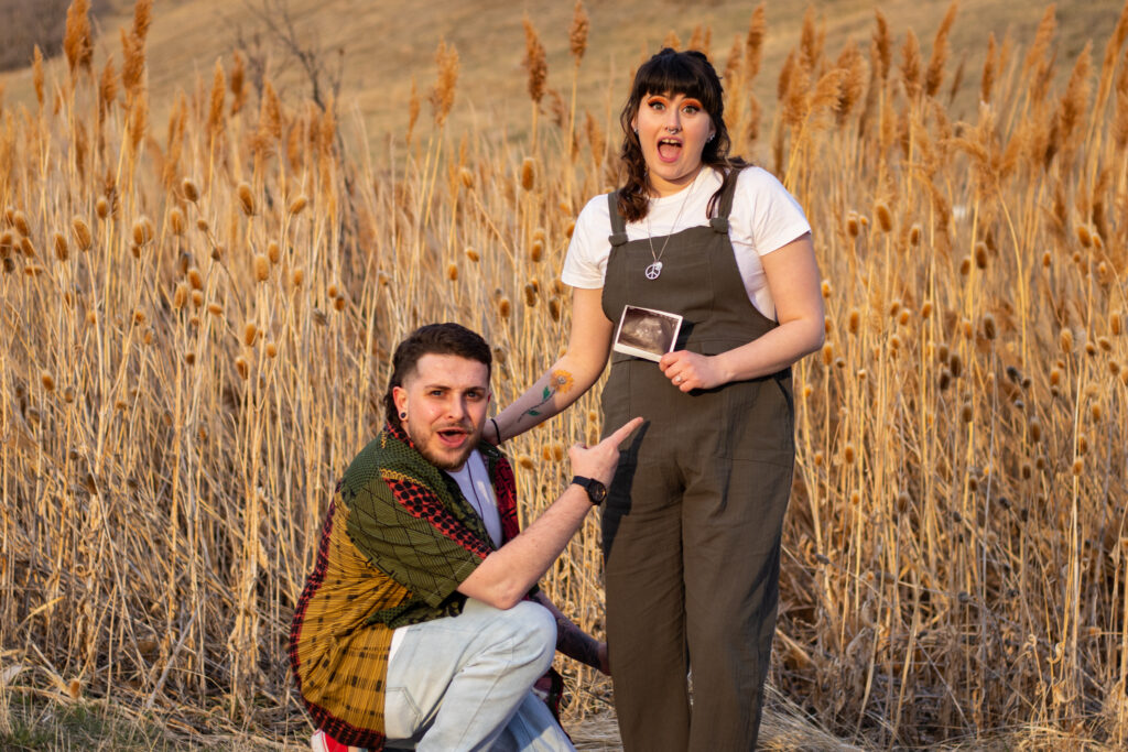 Young couple in a field at Tunnel Springs Park in North Salt Lake. The woman is holding an ultrasound photo standing next to her husband who is crouched down pointing to her belly.